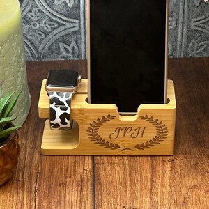 May include: A wooden phone and watch stand with a personalised inscription "JPH" and a decorative laurel wreath design. The stand is made of light brown wood and has a slot for a phone and a watch.