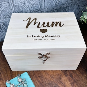 May include: A white wooden box with a hinged lid and a silver latch. The box is engraved with the words "Mum" in cursive, a heart, and the text "In Loving Memory 15 9 1941 - 15/11/2020".