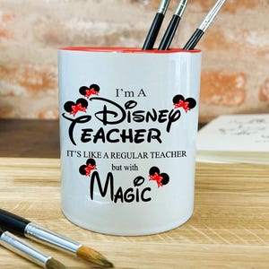 May include: White ceramic mug with a red rim. It features a black and red Mickey Mouse design and the text "I'm a Disney Teacher. It's like a regular teacher, but with magic."
