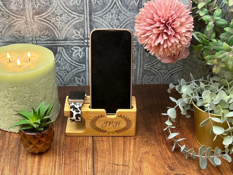 May include: A wooden phone and watch stand with a personalised inscription "JPH" and a decorative laurel wreath design. The stand is made of light brown wood and has a slot for a phone and a watch.