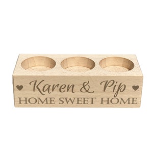May include: A wooden candle holder with three slots for tea lights. It is engraved with the text "Karen & Pip" and "Home Sweet Home" with a heart symbol on each side.
