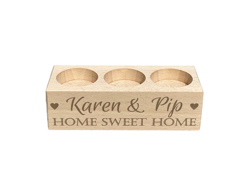 May include: A wooden candle holder with three slots for tea lights. It is engraved with the text "Karen & Pip" and "Home Sweet Home" with a heart symbol on each side.