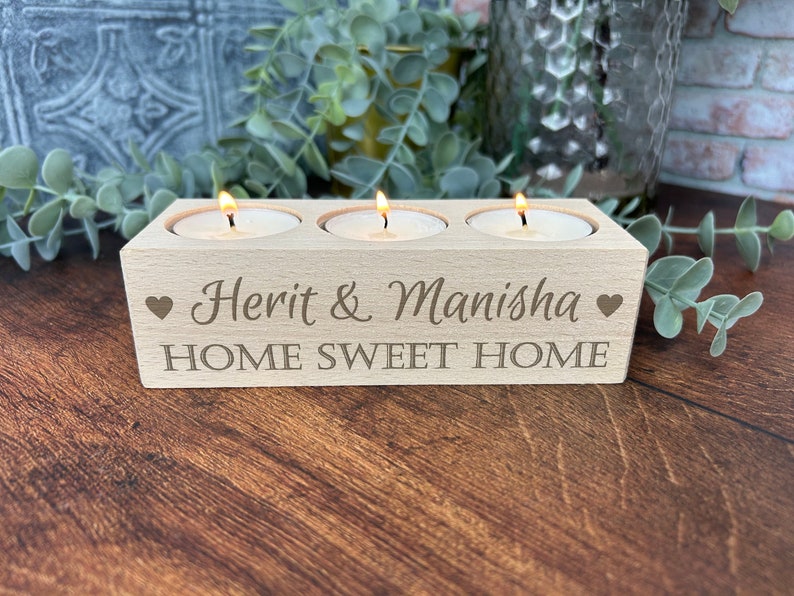 May include: A wooden candle holder with three slots for tea lights. The holder is engraved with the text "Herit & Manisha HOME SWEET HOME" and two hearts.