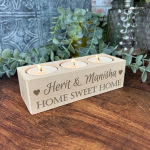 May include: A wooden candle holder with three slots for tea lights. The holder is engraved with the text "Herit & Manisha" and "Home Sweet Home" with a heart symbol on each side.