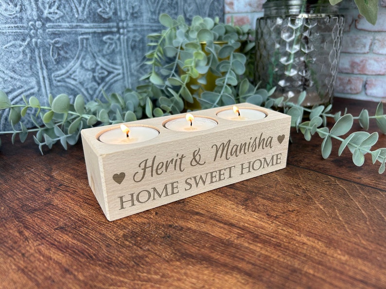May include: A wooden candle holder with three slots for tea lights. The holder is engraved with the text "Herit & Manisha" and "Home Sweet Home" with a heart symbol on each side.