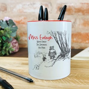 May include: White ceramic pencil holder with a red rim. It features a black and white illustration of Winnie the Pooh and Piglet walking in the woods. The text "Miss Evelleigh Never Forget The Difference You Make" is printed in red on the front.