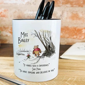 May include: White ceramic pencil holder featuring an illustration of Winnie the Pooh and Piglet. The holder is inscribed with the text "Miss Bailey" and a quote from Winnie the Pooh: "It makes such a difference - Said Pooh - To have someone who believes in you."