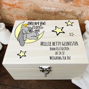 May include: A wooden keepsake box with a light wood finish. The box features a cartoon image of Dumbo the elephant sleeping on a crescent moon with stars. The box is personalised with the text "DREAM BIG little ONE", a name, birthdate, time of birth, and weight.