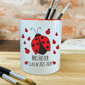 May include: White ceramic pencil holder with a red rim. It features a large ladybug with black spots and red wings, surrounded by smaller ladybugs. The text "Mrs Foster Class of 2023-2024" is printed below the ladybug.