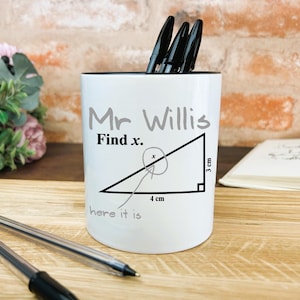 May include: White ceramic pencil holder with the text "Mr Willis Find x." and a triangle with measurements of 3 cm and 4 cm. The holder contains black pens. The background includes a brick wall, flowers, and a notebook.