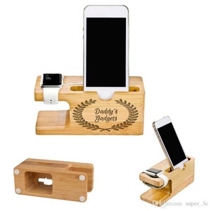 May include: A wooden phone and smartwatch charging stand with a personalised inscription "Daddy's Gadgets" and a laurel wreath design. The stand has a slot for a phone and a watch.