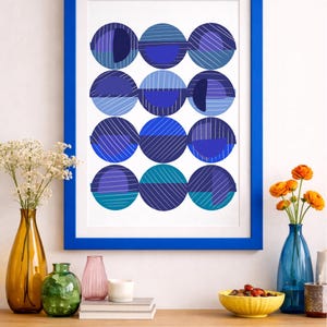 Puede incluir: Una impresión artística enmarcada con un patrón de doce círculos azules con diferentes tonos y líneas diagonales. La obra de arte está en un marco azul. La imagen también muestra jarrones con flores y un cuenco de fruta sobre una superficie de madera.