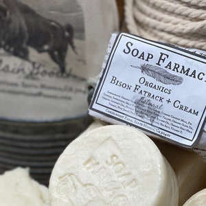 Puede incluir: Una pastilla de jab&oacute;n natural de manteca de bisonte y crema de Soap Farmacy Organics. El jab&oacute;n est&aacute; envuelto en papel blanco con texto negro y un dibujo de una pluma. Los ingredientes est&aacute;n listados en el envoltorio.