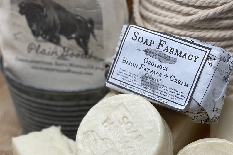 Puede incluir: Una pastilla de jab&oacute;n natural de manteca de bisonte y crema de Soap Farmacy Organics. El jab&oacute;n est&aacute; envuelto en papel blanco con texto negro y un dibujo de una pluma. Los ingredientes est&aacute;n listados en el envoltorio.