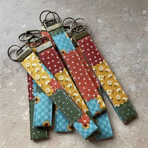 May include: A collection of fabric key fobs with metal key rings. The key fobs feature a patchwork design with floral and geometric patterns in shades of yellow, red, blue, and green. The key rings are a bronze color.