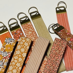 May include: A collection of fabric key fobs in various floral and striped patterns. The key fobs have brass-colored hardware and come in shades of orange, yellow, brown, and beige. The key fobs are arranged on a white surface.