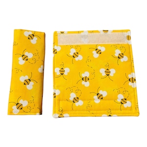 May include: Two yellow fabric items with a repeating pattern of cartoon bees. The bees are black and yellow with white wings. One item is rectangular with a velcro closure, and the other is a smaller, rectangular piece.