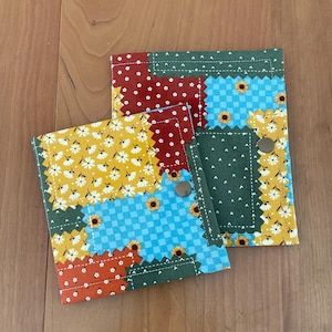 May include: Handmade fabric wallets with a patchwork design. The wallets feature a mix of floral and geometric patterns in red, yellow, blue, and green. Each wallet has a snap closure.