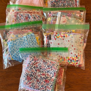 May include: Several clear plastic bags filled with fabric scraps. The fabrics feature floral, polka dot, and patterned designs in various colors, including pink, blue, and red. The bags are sealed with green zippers, and are arranged on a wooden surface.