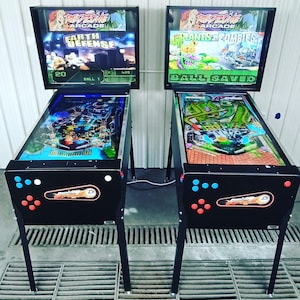 May include: Two black pinball machines with colorful game displays. One reads "Earth Defense" and the other "Plants vs. Zombies." Each machine has a black frame, legs, and control buttons. The machines are set against a white wall.