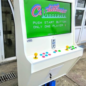 May include: A white arcade machine with a screen displaying the game "Cadillacs and Dinosaurs." The machine has a control panel with joysticks and buttons. The text "PUSH START BUTTON ONLY ONE PLAYER" is visible on the screen.