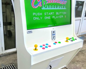 May include: A white arcade machine with a screen displaying the game "Cadillacs and Dinosaurs." The machine has a control panel with joysticks and buttons. The text "PUSH START BUTTON ONLY ONE PLAYER" is visible on the screen.