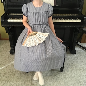 May include: A person wearing a blue and white gingham dress with a white lace fan. The dress has a Peter Pan collar and short sleeves. The person is sitting in front of a black piano.