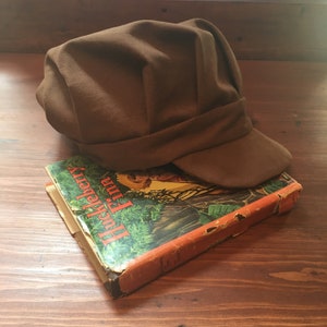 Könnte beinhalten: Eine braune Filz-Newsboy-Mütze mit einem Schirm, die auf einem Buch mit dem Titel "Huckleberry Finn" liegt.