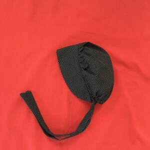 Könnte beinhalten: Eine schwarze und weiße gepunktete Stoffhaube mit einer langen Schleife. Die Haube befindet sich auf einem roten Hintergrund.