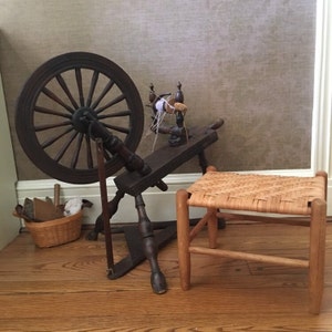 May include: A wooden spinning wheel with a large wooden wheel and a smaller wooden spindle. The spinning wheel is on a wooden stand with four legs. A small wooden stool with a woven seat is in the foreground.