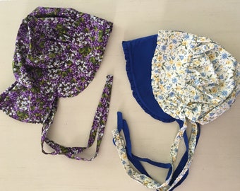 Puede incluir: Dos gorros de bebé. Uno está estampado con flores moradas y blancas, el otro es azul con un estampado floral en amarillo y azul. Ambos tienen lazos para atar debajo de la barbilla.