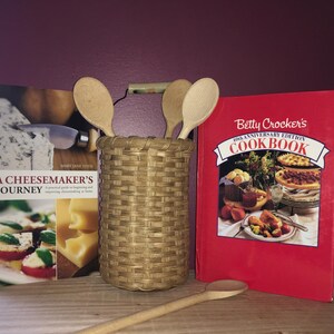 May include: A red cookbook titled "Betty Crocker's 40th Anniversary Edition Cookbook" with a picture of a dessert on the cover.  Two cookbooks, a brown wicker utensil holder, and wooden spoons are on a wooden surface.