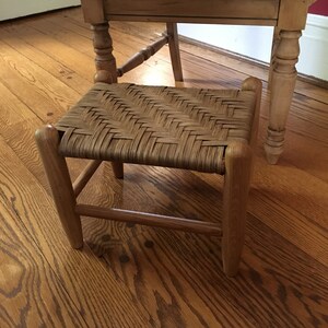 May include: A wooden stool with a woven rush seat. The stool has a simple design with four legs and a rectangular top. The stool is made of natural wood and has a rustic look.