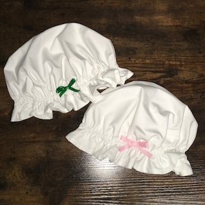 Pode incluir: Dois bonés de tecido branco com bordas franzidas. Um boné tem um laço verde e o outro tem um laço rosa.
