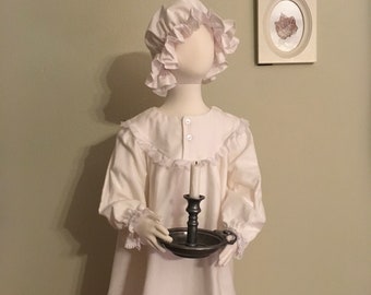 Puede incluir: Un camisón blanco con ribete de encaje y una cofia blanca con volantes. El camisón lo lleva un maniquí que sostiene un candelero con una vela encendida.