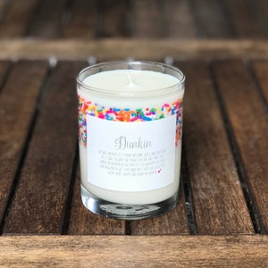 May include: A white candle in a clear glass jar with a colorful sprinkle design. The jar has a label that reads "Dunkin'" and a message about never letting someone become just another day.