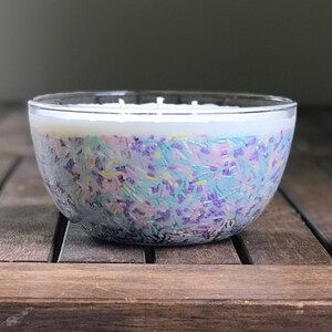 以下が含まれることがあります： 白い上層とカラフルなスプリンクルが入った透明なガラスボウルキャンドル。スプリンクルは青、紫、黄色の色合いです。キャンドルは木製の表面に置かれています。