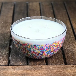 以下が含まれることがあります： 白いワックスの内部と3つの芯を備えた、透明なガラスボウルキャンドル。ボウルの下半分はカラフルなスプリンクルで装飾されています。キャンドルは木製の表面に置かれています。