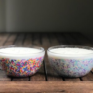 以下が含まれることがあります： 白いワックスとカラフルなスプリンクルが入った、透明なガラスボウルキャンドルが2つ。1つのボウルには赤、黄、青、緑のスプリンクル、もう1つのボウルには紫、青、緑のスプリンクルが入っています。キャンドルは木製の表面に置かれています。