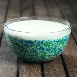 以下が含まれることがあります： 3本の芯が入った、透明なガラスボウルキャンドル。キャンドルのワックスは白で、下半分は青と緑のスプリンクルで飾られています。キャンドルは木製の表面に置かれています。