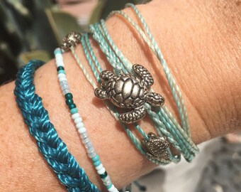 May include: A collection of turquoise and blue bracelets. One bracelet is braided, another has small beads, and the others are thin strands. A silver turtle charm adorns one bracelet. The text "Barrier Island Designs" is in the upper left.