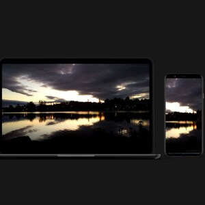 Könnte beinhalten: Ein Laptop und ein Smartphone, die beide dasselbe Bild eines Sees bei Sonnenuntergang mit dunklen Wolken am Himmel zeigen. Das Bild zeigt eine Reflexion des Sonnenuntergangs im Wasser.