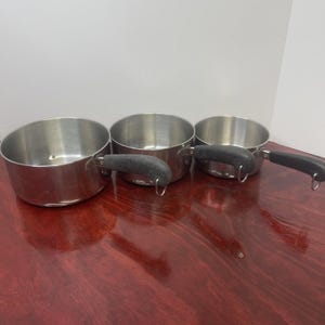 May include: Three stainless steel saucepans of varying sizes arranged in a row. Each pan features a polished silver-tone metal body and a black, textured handle with a small metal loop at the end for hanging. The interiors are smooth and metallic.