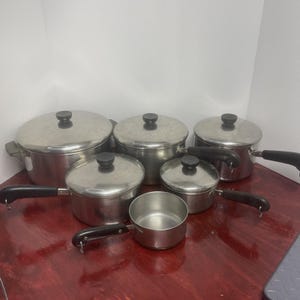Pode incluir: Um conjunto de seis panelas e tachos de aço inoxidável de vários tamanhos. Cada peça apresenta um acabamento prateado polido, uma pega preta e uma tampa a condizer com um botão preto, exceto o pequeno tacho aberto na parte da frente.