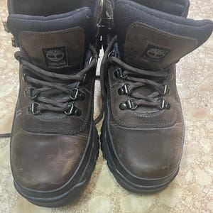 Puede incluir: Un par de botas Timberland de cuero marrón oscuro con diseño de caña alta, cuellos acolchados negros, cordones marrón oscuro, ojales metálicos y suelas gruesas de goma negra. La lengüeta de cada bota muestra un parche con el logo Timberland.
