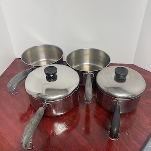 May include: A collection of four stainless steel saucepans with silver-toned metal bodies and black handles. Two of the saucepans feature matching stainless steel lids with black circular knobs, while the other two saucepans remain open.