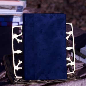 Puede incluir: Un libro azul oscuro se encuentra en posición vertical en un sujetalibros decorativo de color dorado. El libro tiene una cubierta texturizada. Otros libros son visibles en el fondo, junto con una tela drapeada estampada.