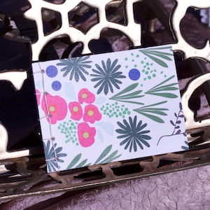 Puede incluir: Un pequeño cuaderno con un estampado floral en rosa, verde y azul descansa sobre un soporte decorativo de metal dorado. La portada del cuaderno presenta un patrón repetido de flores y hojas.