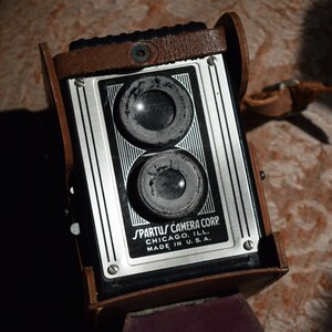 Könnte beinhalten: Eine Vintage-Kamera aus braunem Leder mit zwei runden Objektiven und dem Text "SPARTUS CAMERA CORP. CHICAGO, ILL. MADE IN U.S.A." auf der Vorderseite.