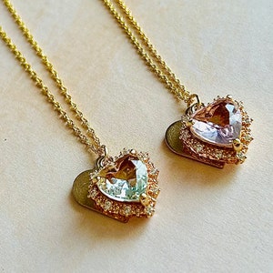 Puede incluir: Dos collares de oro con colgantes en forma de corazón. Los colgantes están adornados con un borde de pequeñas piedras transparentes y presentan una piedra facetada más grande en el centro. Un colgante tiene una piedra rosa claro y el otro una piedra azul claro.